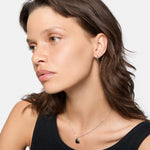 Frau mit gewellten braunen Haaren in einem schwarzen Oberteil, trägt silberne LIEBESKIND BERLIN Creole - Urban Heart Ohrringe und eine Liebeskind Berlin Herzanhänger Halskette, schaut nach links.