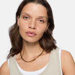 Eine Frau mit gewellten braunen Haaren trägt die LIEBESKIND BERLIN Halskette - Urban Chic von Liebeskind Berlin, goldene Ohrringe und ein beiges Tank-Top über Weiß - der perfekte Look für Statement-Schmuck-Liebhaberinnen.