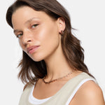 Frau mit gewellten braunen Haaren trägt ein beiges Tanktop, goldene Ohrringe und die LIEBESKIND BERLIN Halskette - Urban Chic von Liebeskind Berlin.