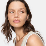 Frau mit gewellten braunen Haaren in einem beigen Oberteil modelliert die LIEBESKIND BERLIN Halskette - Urban Chic von Liebeskind Berlin und blickt nach links auf einen weißen Hintergrund.