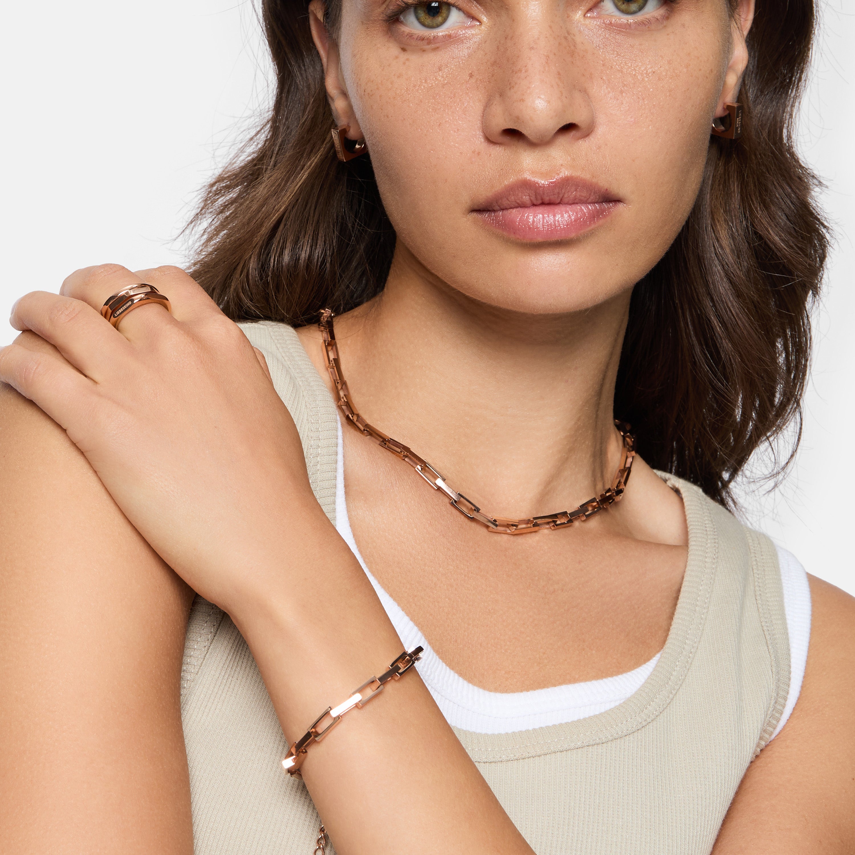 Frau in einem ärmellosen beigen Top trägt roségoldenen Schmuck, darunter den LIEBESKIND BERLIN Ring - Urban Chic in 54, zusammen mit passender Halskette, Armband und Ohrringen von Liebeskind Berlin.