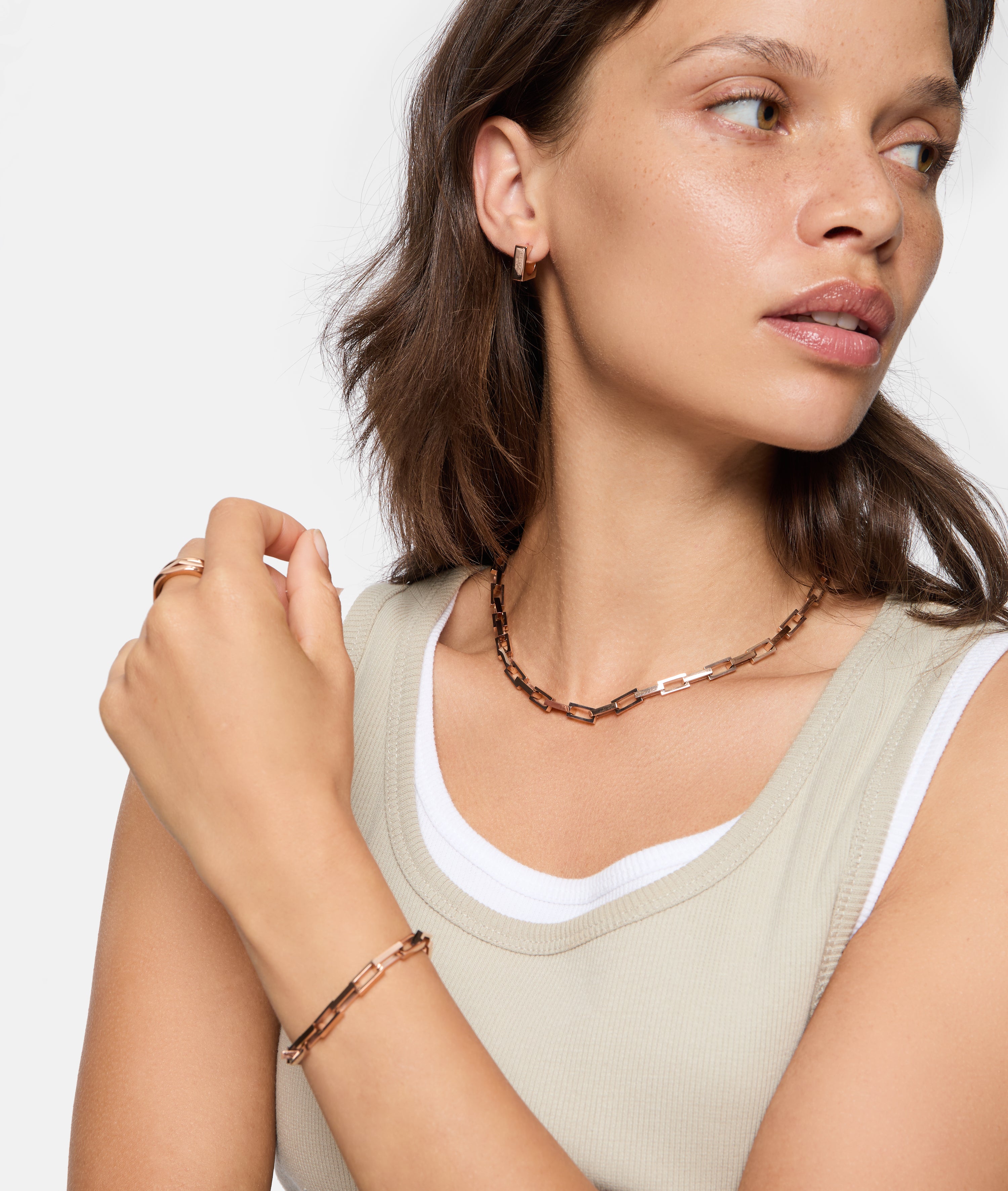 Frau trägt LIEBESKIND BERLIN Halskette - Urban Chic, goldenes Armband, Ring und Ohrringe von Liebeskind Berlin, kombiniert mit einem beigefarbenen ärmellosen Oberteil.