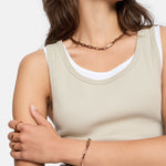Die Frau im beigen Tank-Top trägt eine goldene Halskette, ein Armband, Ringe und das LIEBESKIND BERLIN Armband - Urban Chic von Liebeskind Berlin und kreuzt ihren Arm über der Brust für einen modernen urbanen Look.