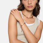 Eine Frau in einem beigen Tanktop trägt goldenen Schmuck, darunter das Liebeskind Berlin Urban Chic Armband, eine Halskette, einen Ring und kleine Ohrringe für einen urbanen Look.