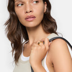 Eine Frau mit gewellten braunen Haaren in einem Tanktop und Goldschmuck trägt den LIEBESKIND BERLIN Ring - Urban Chic in 54 von Liebeskind Berlin und hält ein schwarzes Armband über ihre Schulter.