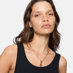 Eine Frau mit schulterlangen braunen Haaren trägt ein schwarzes Tanktop und die Liebeskind Berlin Halskette "The T-Bar Plate" mit einem goldenen Epoxy-Anhänger und einem stilvollen T-Bar-Verschluss.