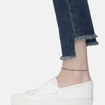 Eine Person trägt eine Jeans mit ausgefranstem Saum, weiße Slipper und die LIEBESKIND BERLIN Fußkette - The Simple Solo von Liebeskind Berlin vor einem schlichten Hintergrund.