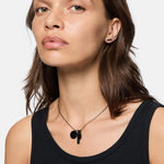 Frau mit braunen Haaren trägt ein schwarzes Tanktop, schwarze Ohrringe und die Liebeskind Berlin Halskette - The Talisman und schaut in die Kamera.