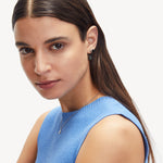 Frau mit glattem braunem Haar trägt ein blaues ärmelloses Oberteil, Liebeskind Berlin Halskette und LIEBESKIND BERLIN Ohrstecker - The Knot Ohrringe von Liebeskind Berlin und schaut in die Kamera.