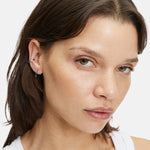 Eine Frau mit heller Haut und braunen Haaren trägt The Organic Creole Ohrringe von Liebeskind Berlin und ein weißes Tanktop und schaut in die Kamera.