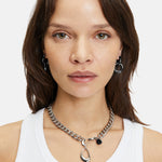 Frau mit glattem braunem Haar trägt ein weißes Tank-Top und Liebeskind Berlin-Schmuck, darunter The Organic Creole-Ohrringe (LIEBESKIND BERLIN Creole) und eine Halskette mit abnehmbarem Anhänger, mit Blick in die Kamera.