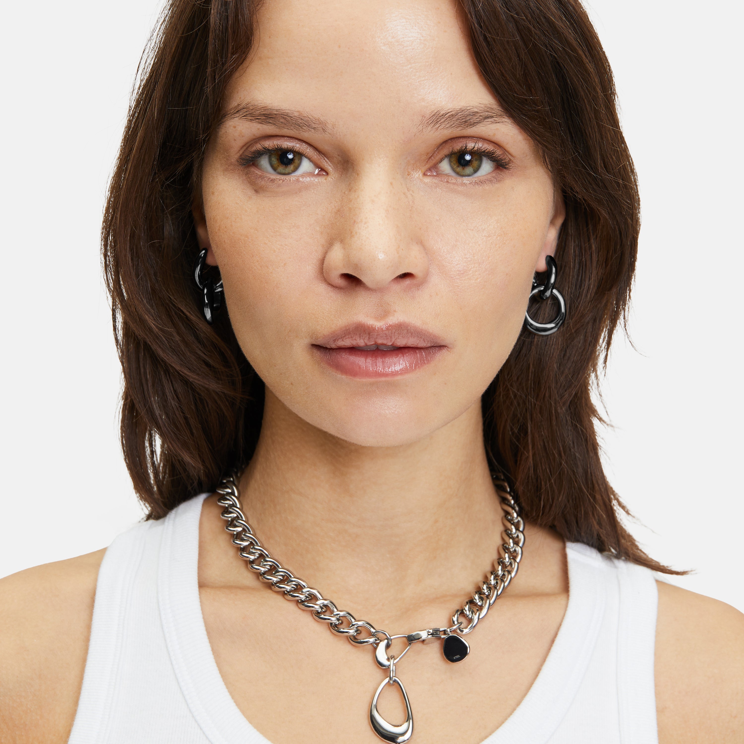 Frau mit glattem braunem Haar trägt ein weißes Tank-Top und Liebeskind Berlin-Schmuck, darunter The Organic Creole-Ohrringe (LIEBESKIND BERLIN Creole) und eine Halskette mit abnehmbarem Anhänger, mit Blick in die Kamera.