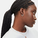 Frau mit geflochtenem Pferdeschwanz, in weißem Hemd und Ohrring, trägt eine Liebeskind Berlin Halskette - 3D Lettering mit rundem Anhänger, von rechts.