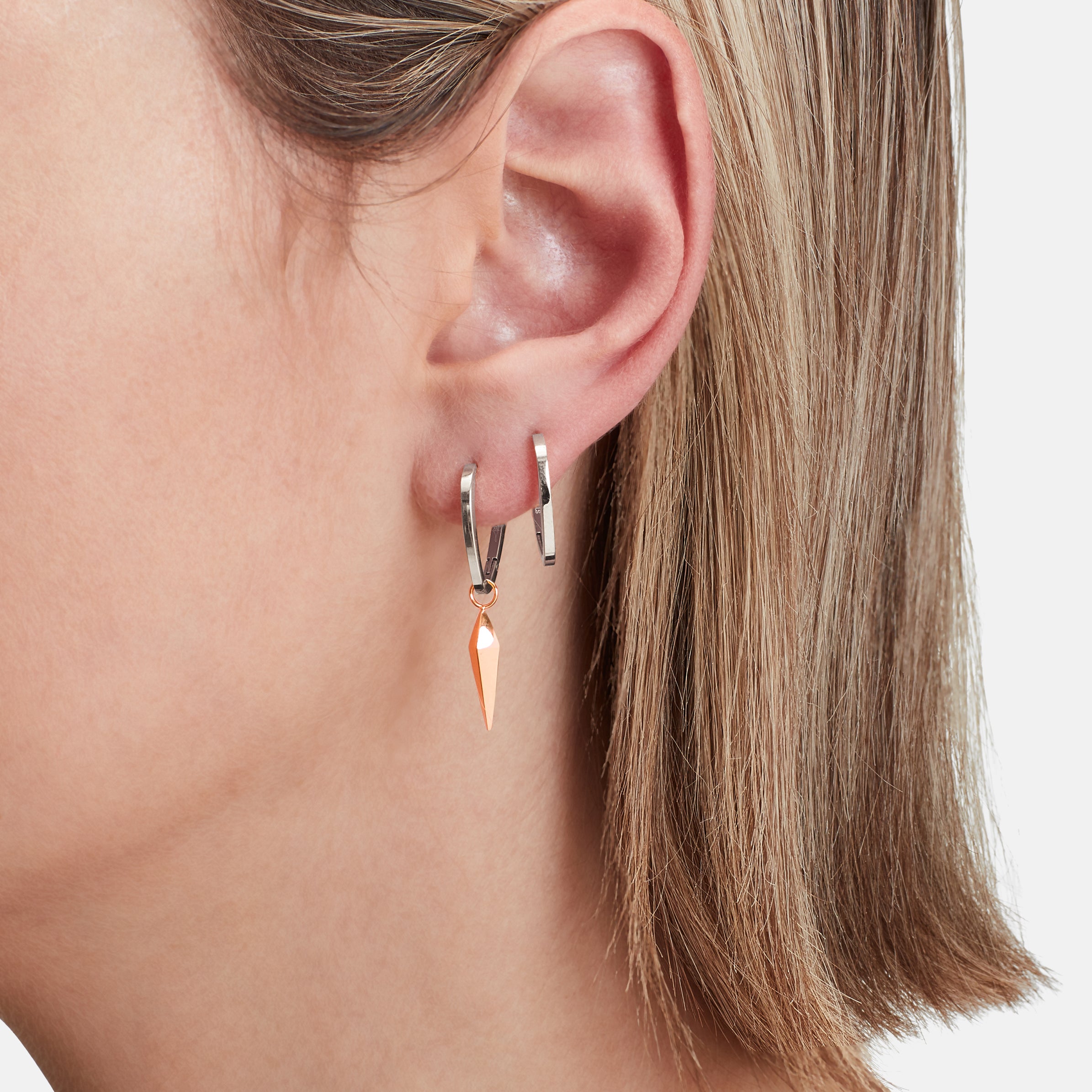 Nahaufnahme des Ohres einer Person mit zwei silbernen Creolen, eine davon mit einem spitzen orangefarbenen Anhänger. Entdecken Sie LIEBESKIND BERLIN Creole - The Pendulum, stilvoller Trendschmuck von Liebeskind Berlin.