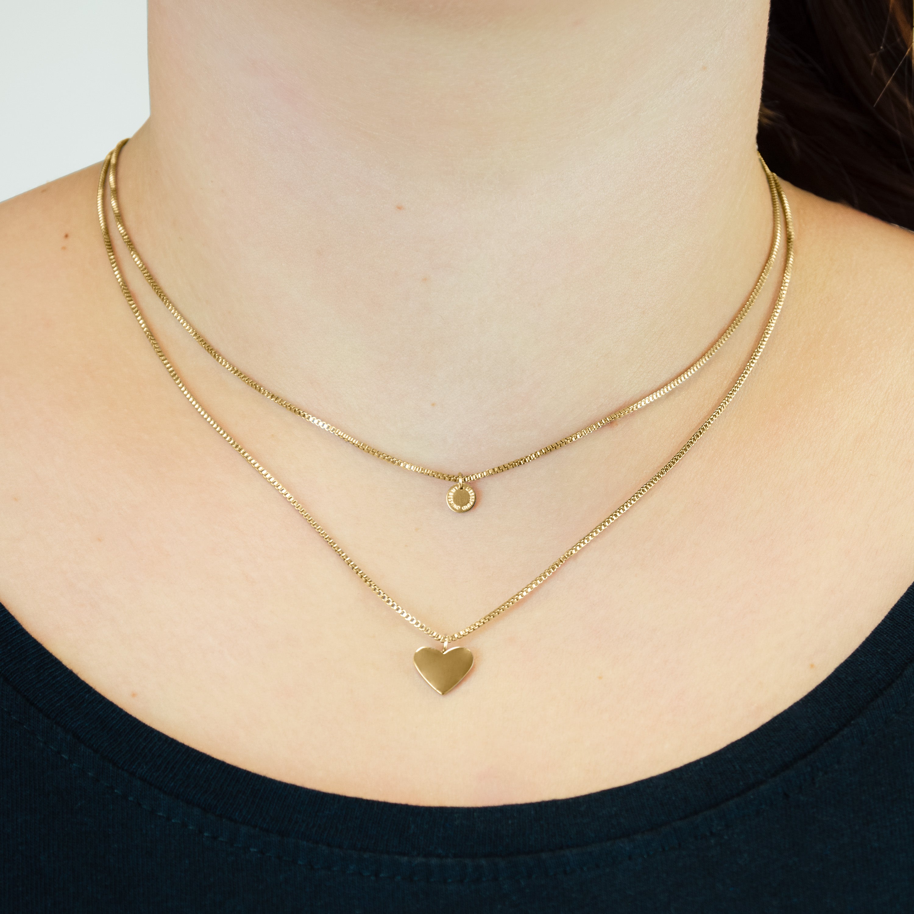 Eine Person trägt die Liebeskind Berlin Halskette - Layered Love: zwei goldene Halsketten, eine mit einem kleinen Kreisanhänger und die andere mit einem Herzanhänger.