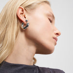 Blonde Frau in einem grauen Oberteil, die die Ohrringe Steel Ball Creole von Liebeskind Berlin trägt, mit geneigtem Kopf und geschlossenen Augen.