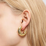 Nahaufnahme eines Ohrs mit blondem Haar, das klobige goldene LIEBESKIND BERLIN Creole - Steel Ball Ohrringe von Liebeskind Berlin und zwei kleine goldene Ohrringe trägt.