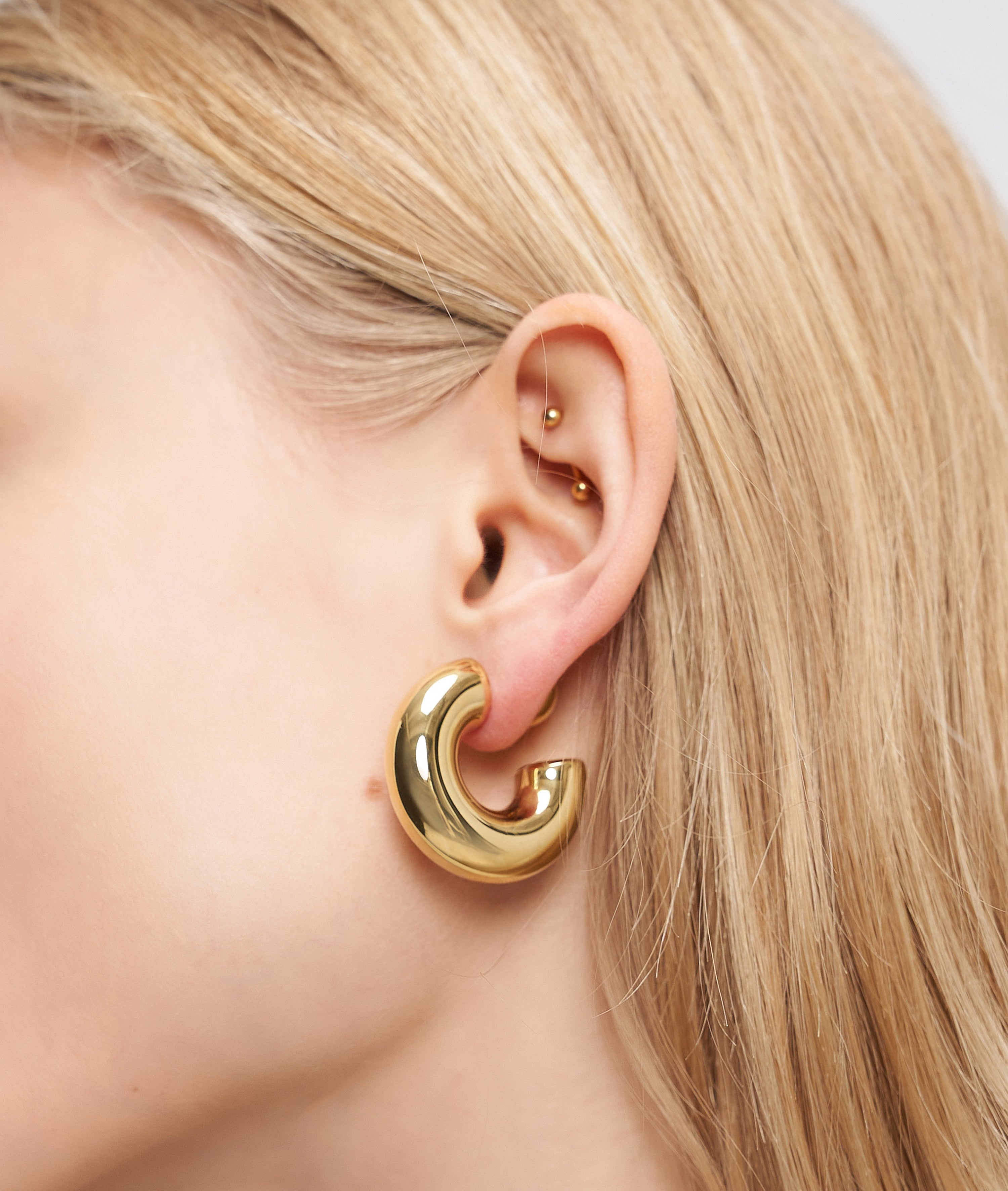 Nahaufnahme eines Ohrs mit blondem Haar, das klobige goldene LIEBESKIND BERLIN Creole - Steel Ball Ohrringe von Liebeskind Berlin und zwei kleine goldene Ohrringe trägt.