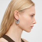 Frau mit langen blonden Haaren im Seitenprofil trägt die LIEBESKIND BERLIN Creole - Organic Shapes Ohrringe von Liebeskind Berlin und ein braunes Stricktop - ein perfektes Beispiel für minimalistischen Schmuck.