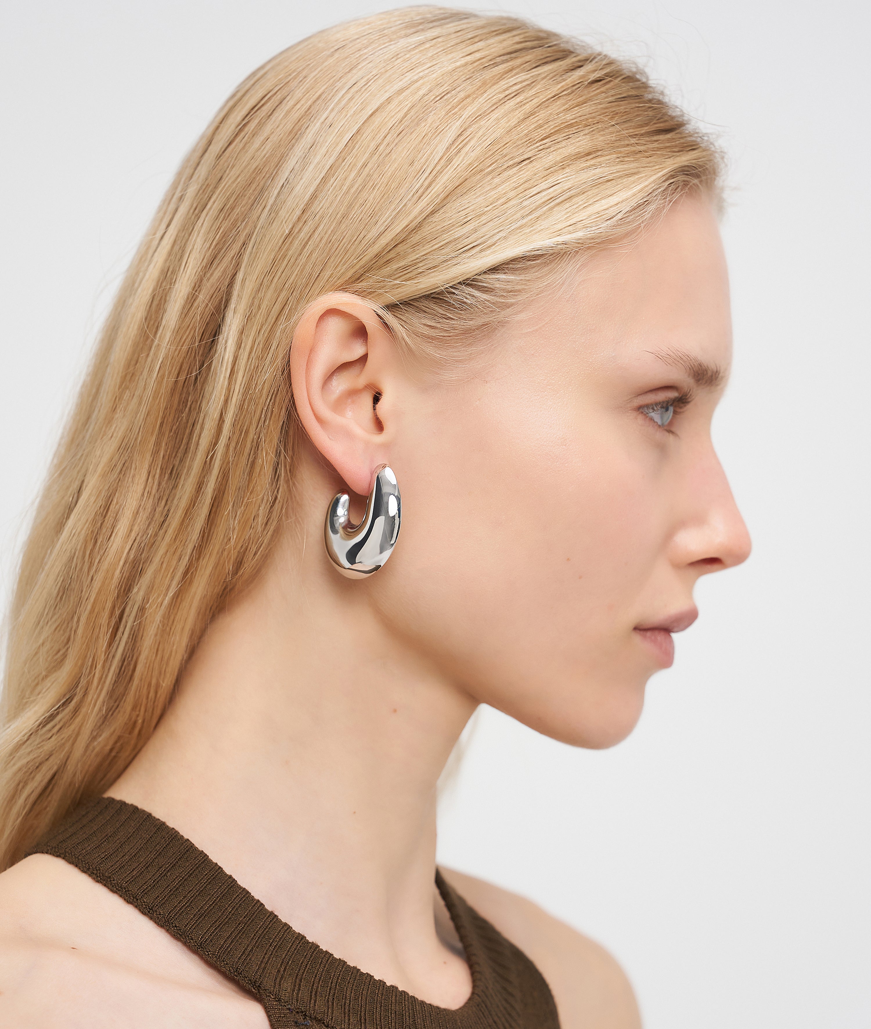Frau mit langen blonden Haaren im Seitenprofil trägt die LIEBESKIND BERLIN Creole - Organic Shapes Ohrringe von Liebeskind Berlin und ein braunes Stricktop - ein perfektes Beispiel für minimalistischen Schmuck.