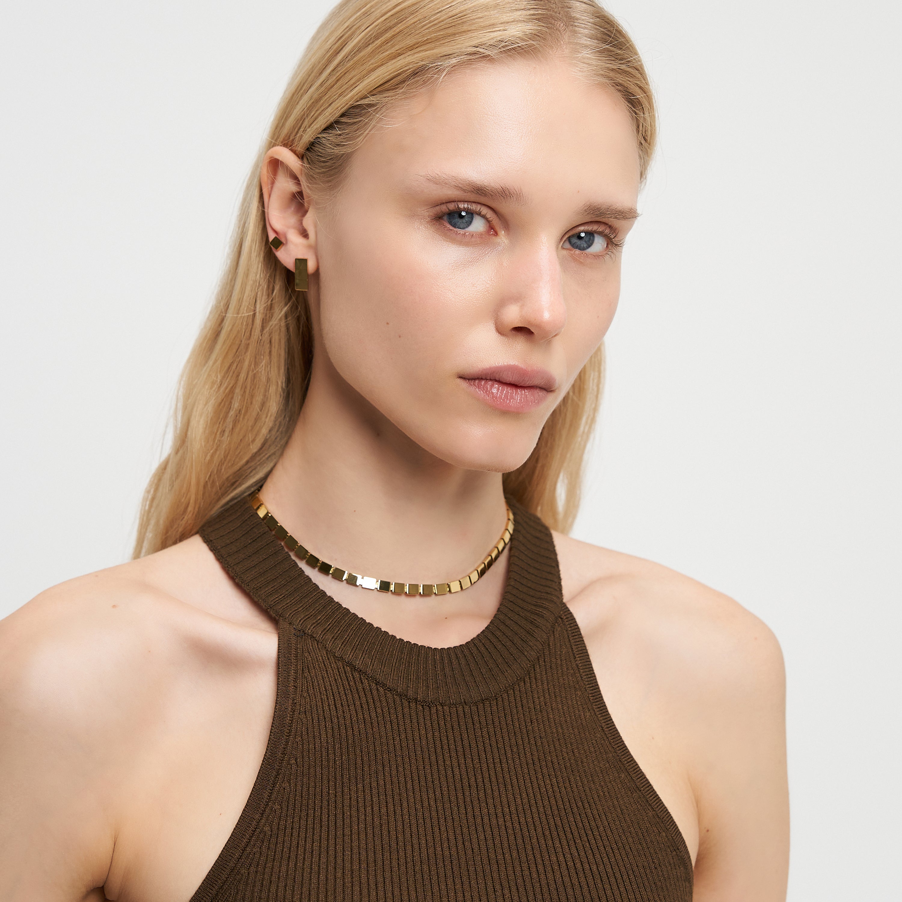 Frau mit langen blonden Haaren, die ein braunes Neckholder-Top, Goldschmuck und die LIEBESKIND BERLIN Halskette - Plates by Liebeskind Berlin trägt, schaut in die Kamera.
