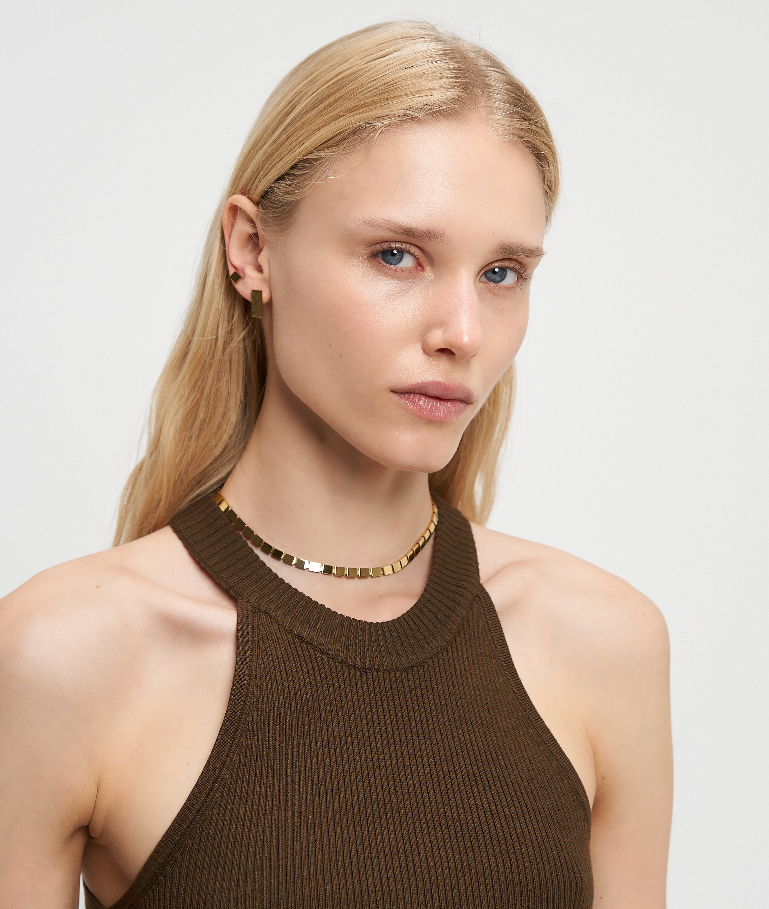 Frau mit langen blonden Haaren, die ein braunes Neckholder-Top, Goldschmuck und die LIEBESKIND BERLIN Halskette - Plates by Liebeskind Berlin trägt, schaut in die Kamera.