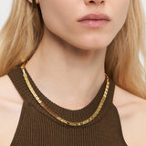 Frau trägt eine LIEBESKIND BERLIN Halskette - Plates von Liebeskind Berlin, gepaart mit goldenen rechteckigen Ohrringen und einem braunen gerippten Top für einen eleganten Damen Accessoire Look.
