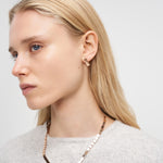 Frau mit langen blonden Haaren trägt die Liebeskind Berlin Halskette - Plates, kleine Ohrringe und ein hellgraues Oberteil und blickt zur Seite.