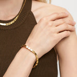 Eine Frau in einem ärmellosen braunen Ripp-Top berührt ihre Schulter, gestylt mit dem goldenen LIEBESKIND BERLIN Armband - Plates von Liebeskind Berlin und einer passenden Halskette - ideal für einen urbanen Look.