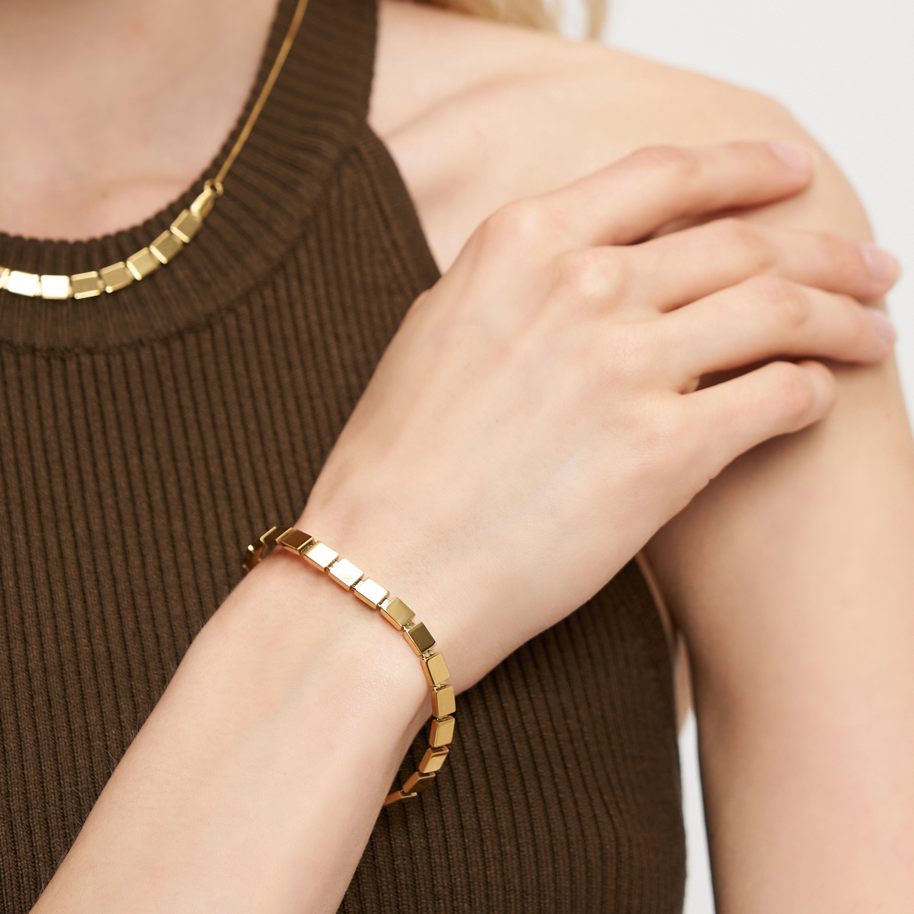 Eine Frau in einem ärmellosen braunen Ripp-Top berührt ihre Schulter, gestylt mit dem goldenen LIEBESKIND BERLIN Armband - Plates von Liebeskind Berlin und einer passenden Halskette - ideal für einen urbanen Look.