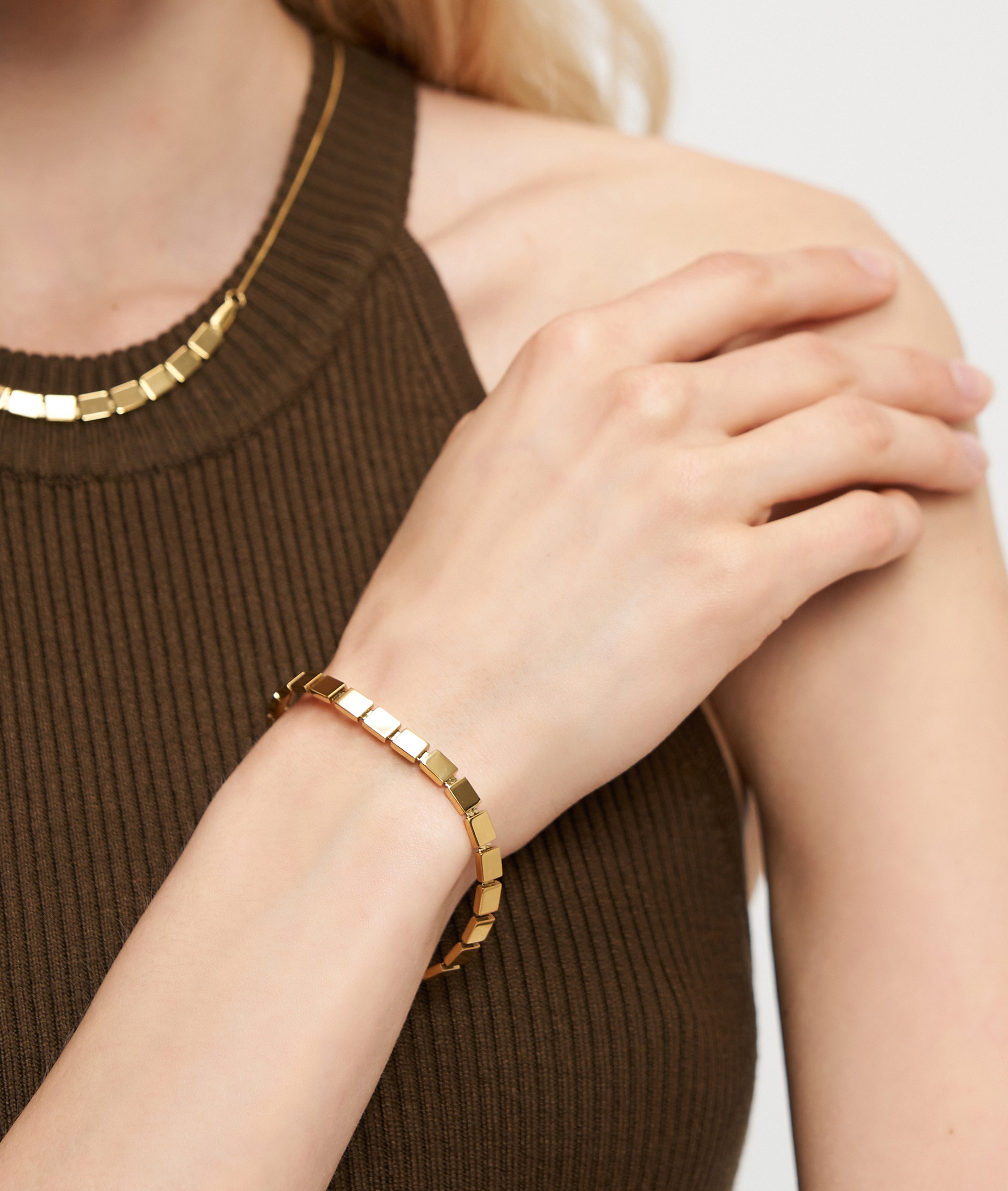 Eine Frau in einem ärmellosen braunen Ripp-Top berührt ihre Schulter, gestylt mit dem goldenen LIEBESKIND BERLIN Armband - Plates von Liebeskind Berlin und einer passenden Halskette - ideal für einen urbanen Look.