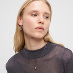 Eine Frau mit langen blonden Haaren trägt ein durchsichtiges graues Oberteil, silberne Liebeskind Berlin Creole - Glass Heart Ohrringe und eine passende Halskette mit Herzanhänger.