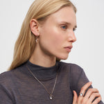 Eine Frau mit langen blonden Haaren trägt Liebeskind Berlin Creole - Glass Heart Ohrringe, eine Halskette mit Herzanhänger und einen Ring, gestylt in einem durchsichtigen dunklen Top.