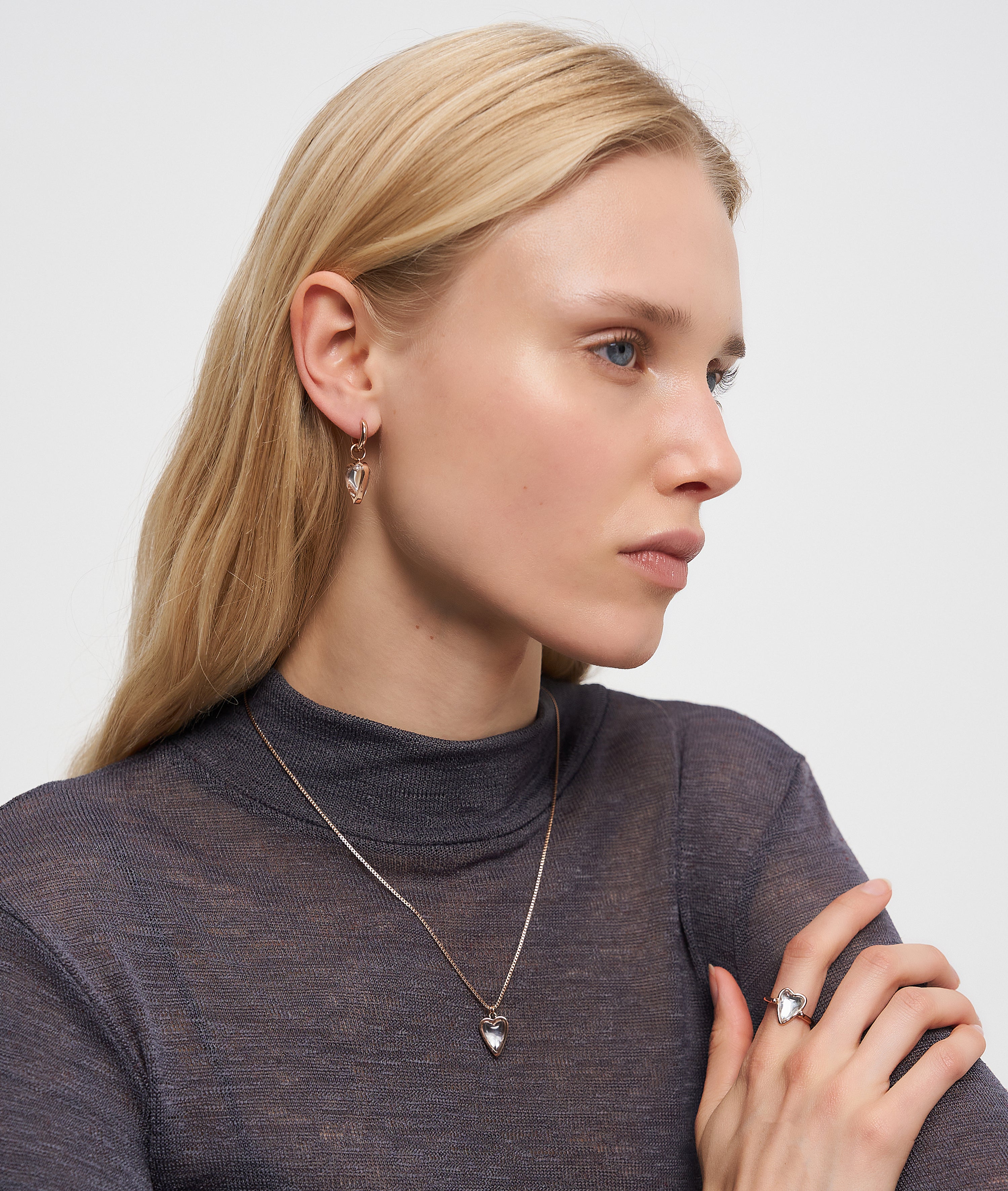 Eine Frau mit langen blonden Haaren trägt Liebeskind Berlin Creole - Glass Heart Ohrringe, eine Halskette mit Herzanhänger und einen Ring, gestylt in einem durchsichtigen dunklen Top.