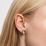 Nahaufnahme eines Frauenohrs, das den Liebeskind Berlin Ohrstecker - Glass Heart trägt, einen goldenen herzförmigen Ohrring mit einem klaren Edelstein.