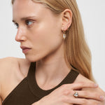 Eine Frau mit langen blonden Haaren trägt ein braunes Neckholder-Top, goldene herzförmige Ohrringe mit zartem Herzdetail und den LIEBESKIND BERLIN Ring - Glass Heart von Liebeskind Berlin als ihr besonderes Schmuckstück.