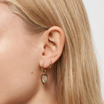 Nahaufnahme eines Frauenohrs mit drei goldenen Ohrringen, darunter die Liebeskind Berlin Creole - Glass Heart, vor einem neutralen Hintergrund.