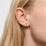 Nahaufnahme eines Ohres mit langen blonden Haaren, das den kleinen, silbernen LIEBESKIND BERLIN Ohrstecker - Glass Heart von Liebeskind Berlin im zarten Herzdesign trägt.