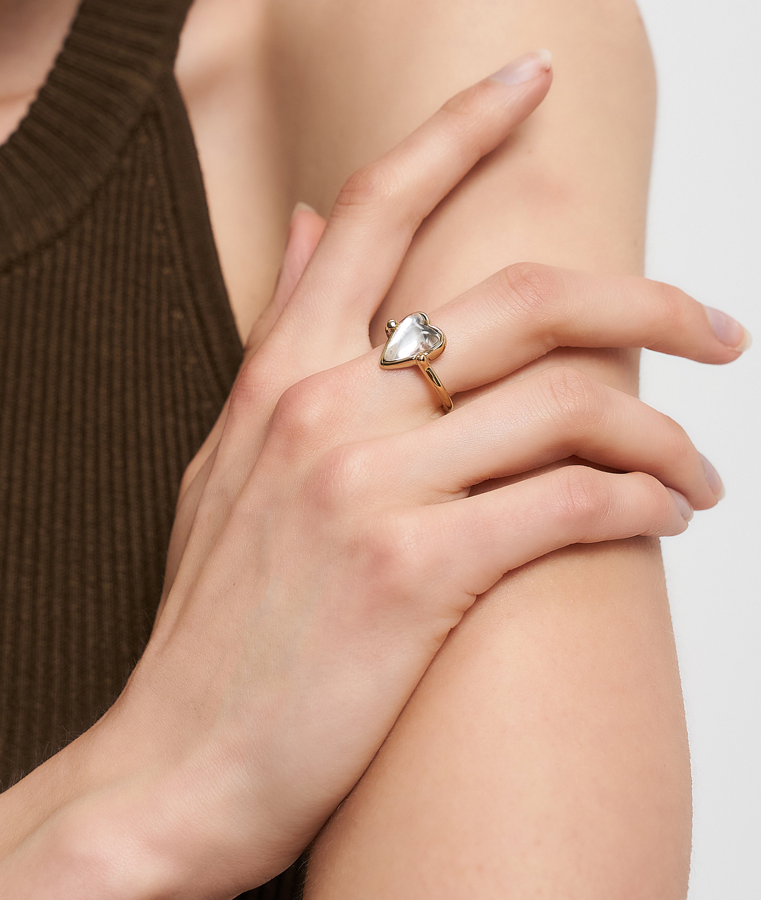 Eine Hand trägt den goldenen Liebeskind Berlin Ring - Glass Heart mit zartem Herzmotiv und Perle, der sanft auf einem nackten Arm ruht - ein elegantes Stück voller Charme.