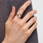 Eine Hand mit dem Liebeskind Berlin Ring - Glass Heart, der ein gläsernes Herz auf einem schlanken Band zeigt, ruht anmutig auf dunklem, strukturiertem Stoff.