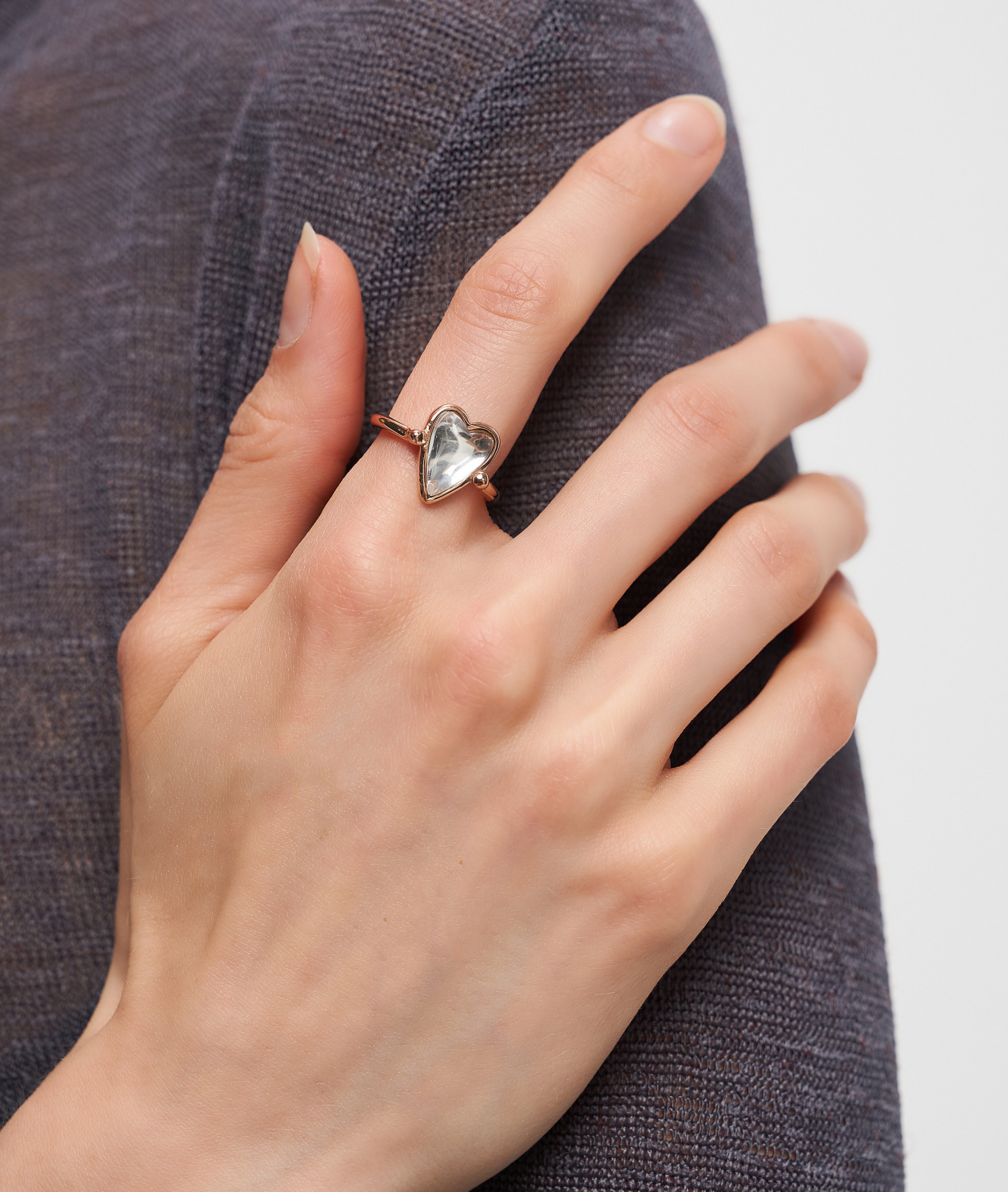 Eine Hand mit dem Liebeskind Berlin Ring - Glass Heart, der ein gläsernes Herz auf einem schlanken Band zeigt, ruht anmutig auf dunklem, strukturiertem Stoff.