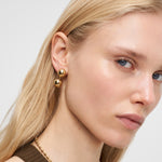 Eine Frau mit glattem blondem Haar trägt Liebeskind Berlin Ohrring - Steel Ball und eine passende goldene Halskette und schaut in die Kamera.