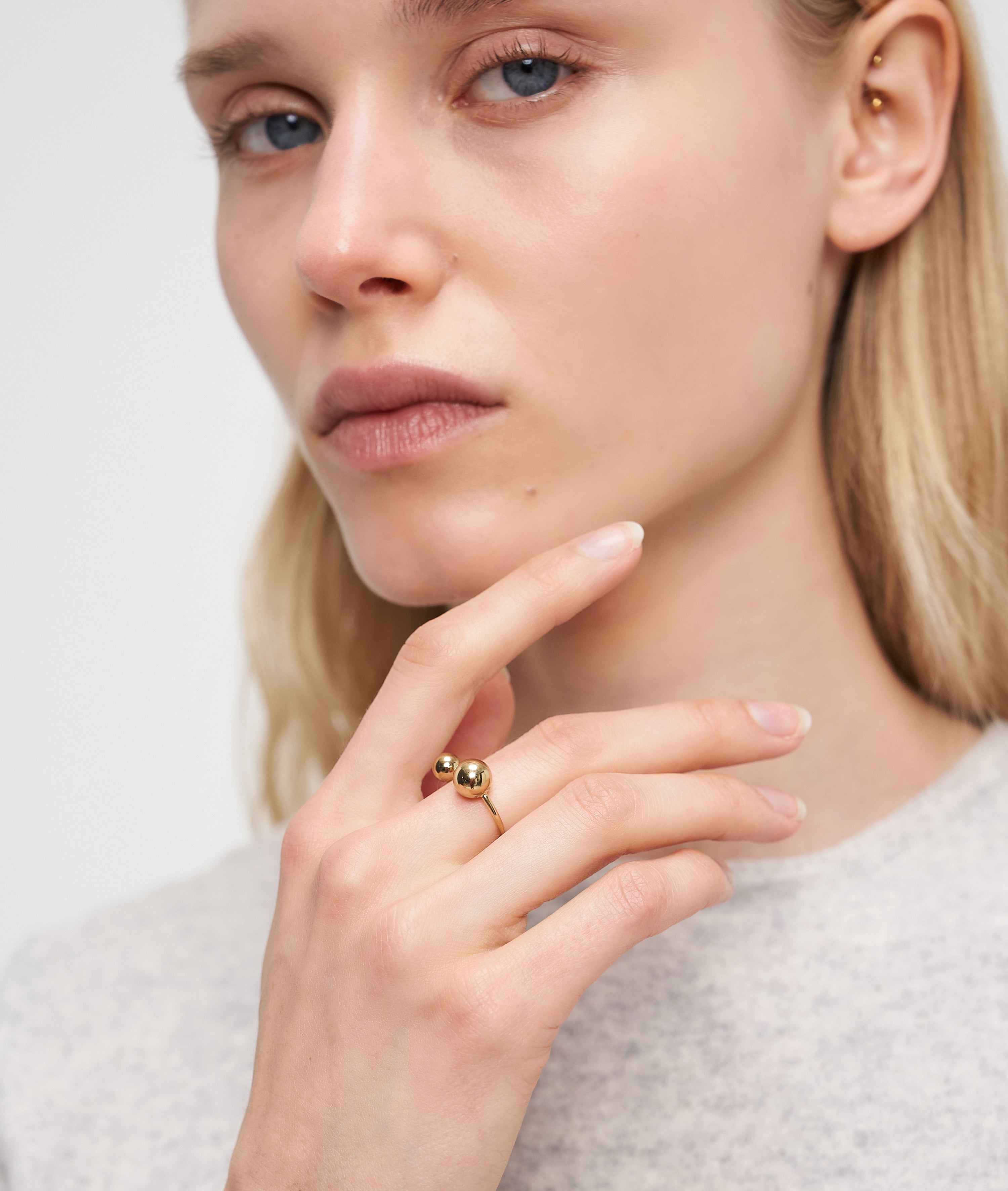 Eine blonde Frau, die einen leichten Pullover und den LIEBESKIND BERLIN Ring - Steel Ball von Liebeskind Berlin trägt, berührt ihr Gesicht und blickt in die Kamera. Sie zeigt mühelosen Stil mit modernem Design.