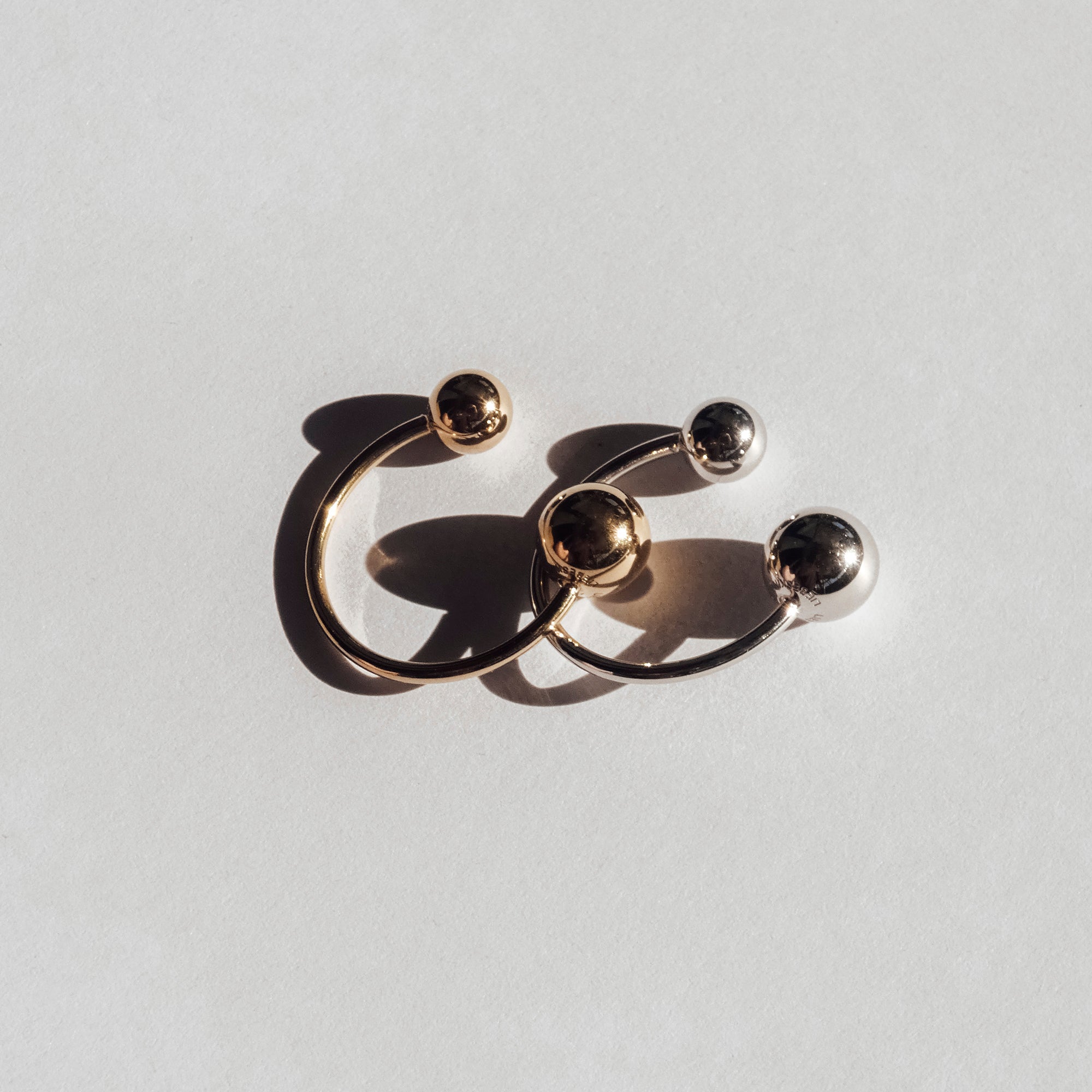Zwei offene Ringe, ein goldener und ein silberner, jeweils mit Stahlkugelenden, präsentieren ein modernes Design aus der LIEBESKIND BERLIN Ring - Steel Ball Kollektion von Liebeskind Berlin auf weißer Oberfläche.