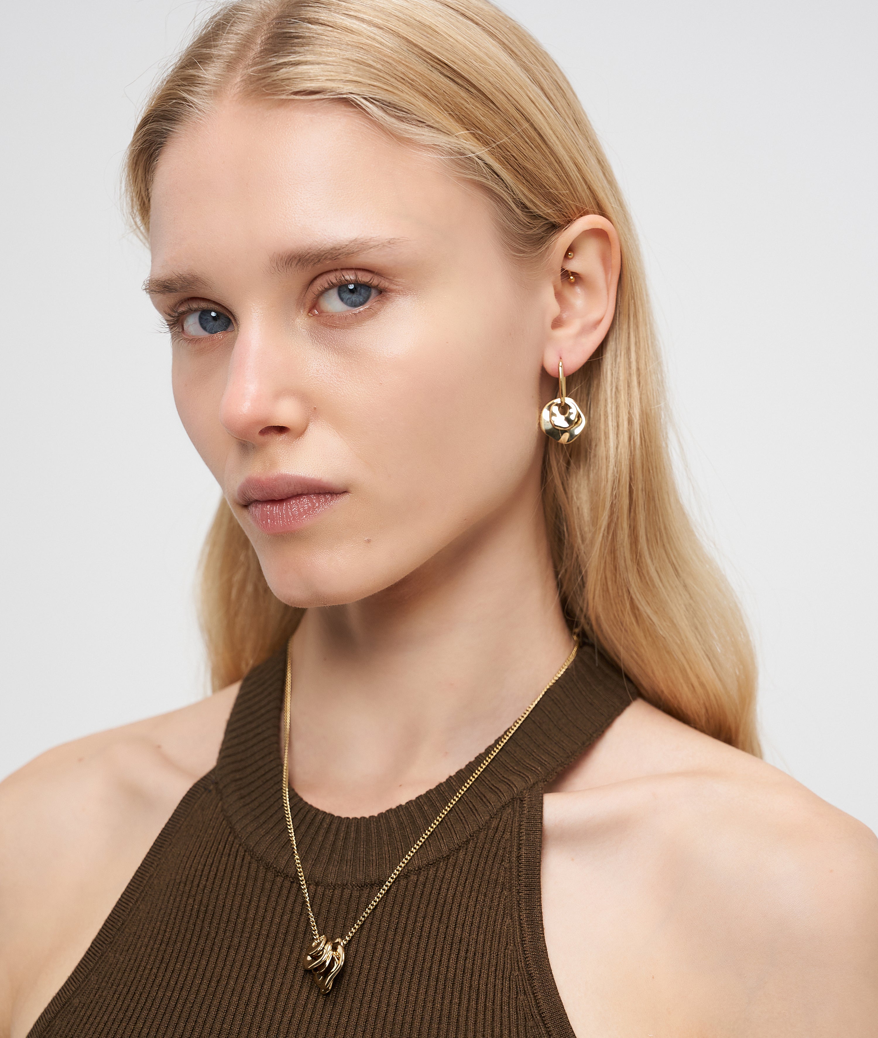 Frau mit langen blonden Haaren trägt eine LIEBESKIND BERLIN Halskette - Organic Shapes von Liebeskind Berlin, gepaart mit goldenem Schmuck, stilvollen goldenen Ohrringen und einem ärmellosen braunen Top.