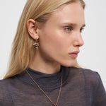 Frau mit langen blonden Haaren, die ein durchsichtiges dunkles Oberteil, Ohrringe und eine Liebeskind Berlin Halskette - Organic Shapes trägt und zur Seite schaut.