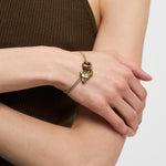 Eine Person, die das goldene LIEBESKIND BERLIN Armband - Organic Shapes von Liebeskind Berlin mit einem Katzenkopf-Anhänger trägt, gepaart mit einem ärmellosen braunen gerippten Top.