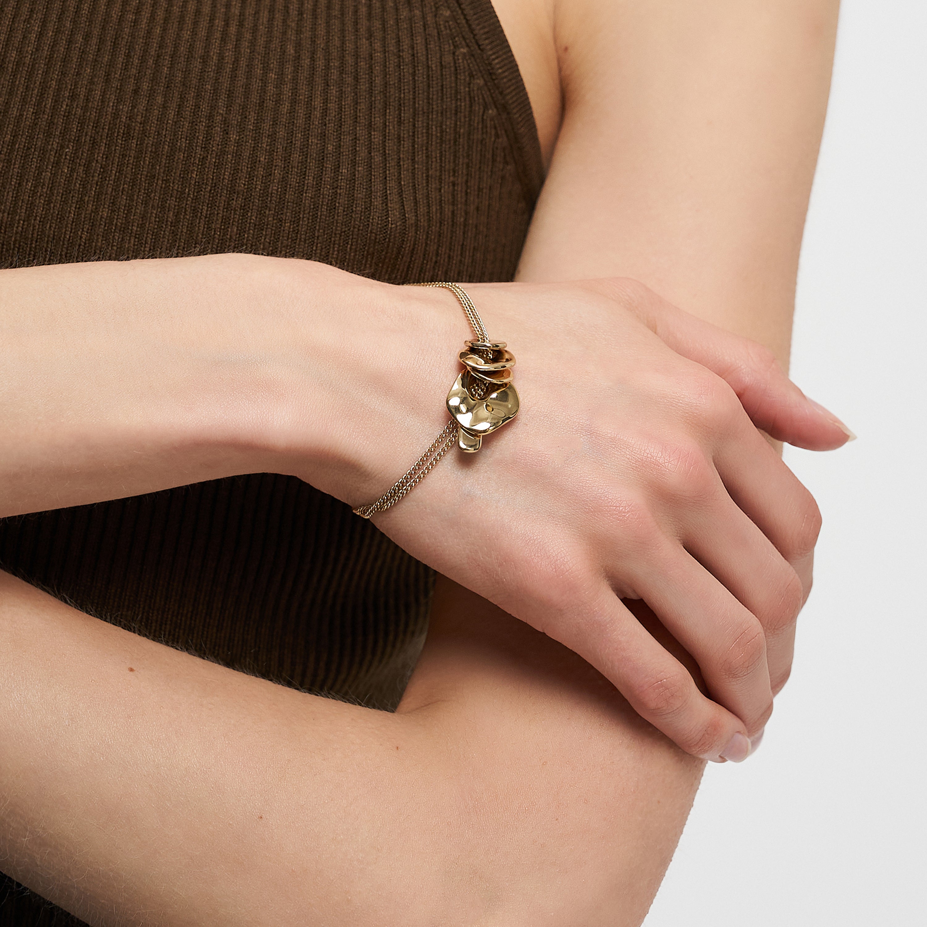 Eine Person, die das goldene LIEBESKIND BERLIN Armband - Organic Shapes von Liebeskind Berlin mit einem Katzenkopf-Anhänger trägt, gepaart mit einem ärmellosen braunen gerippten Top.