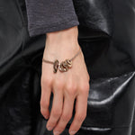 Eine Hand, die das LIEBESKIND BERLIN Armband - Organic Shapes von Liebeskind Berlin mit zarten Kettengliedern und mehreren baumelnden Anhängern trägt, ruht auf einer schwarzen Lederhose.