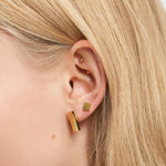 Eine Nahaufnahme des Ohrs einer Frau mit kurzem blondem Haar zeigt den geometrischen goldenen Liebeskind Berlin Ohrring - Plates und ein oberes Ohrpiercing für einen architektonischen Look.